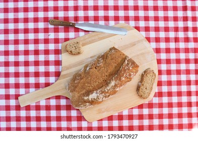 Homemade Whole Wheat Artesian Bread Loaf On A Pizza Peel Board With A Red And White Table Cloth.