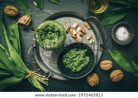 Homemade wild garlic pesto on a metal tray, wild garlic leaves, salt, oil and walnuts on dark green background, top view