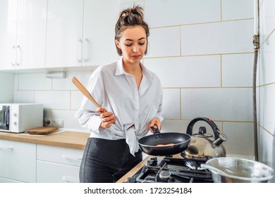 Homemade Woman Likes To Cook Delicious Desserts From Natural Products. Making Banana Pancakes, Turning Over By Wooden Spatula The Already Fried Side Of The Fritters In The Pan. Smiling, Feeling Happy