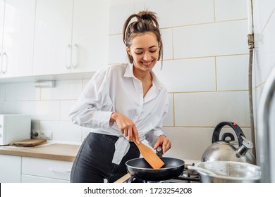 Homemade Woman Likes To Cook Delicious Desserts From Natural Products. Making Banana Pancakes, Turning Over By Wooden Spatula The Already Fried Side Of The Fritters In The Pan. Smiling, Feeling Happy