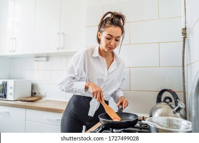 Homemade Woman Likes To Cook Delicious Desserts From Natural Products. Making Banana Pancakes, Turning Over By Wooden Spatula The Already Fried Side Of The Fritters In The Pan. Smiling, Feeling Happy