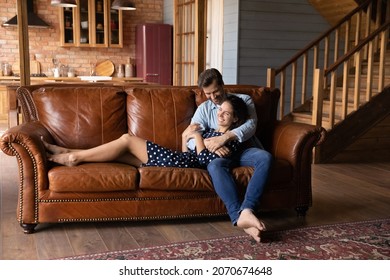 Homeowners. Happy Millennial Spouses Buyers Renters Of New House Rest Barefoot On Modern Couch At Wooden Cottage Interior. Affectionate Young Couple Husband Wife Hug On Cozy Sofa Spend Weekend At Home