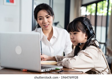 Homeschool Asian Little Young Girl Learning Online Class From School Teacher By Digital Remote Internet Meeting Due To Coronavirus Pandemic. Kid Looking Computer And Writing Note, Sitting With Mother.