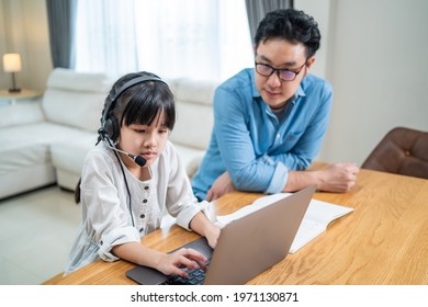 Homeschool Asian Little Young Girl Learning Online Class From School Teacher By Digital Remote Internet Meeting Due To Coronavirus Pandemic. Kid Looking Computer And Writing Note With Help From Father