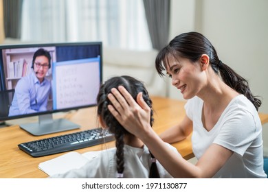 Homeschool Asian little young girl learning online class from school teacher by digital remote internet meeting due to coronavirus pandemic. Kid looking computer and writing note with help from mother