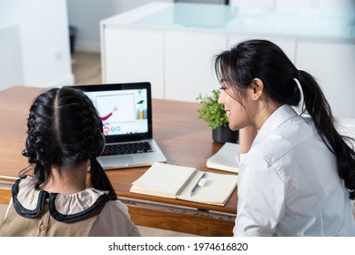 Homeschool Asian Little Young Girl Learning Online Class From School Teacher By Digital Remote Internet Meeting Due To Coronavirus Pandemic. Kid Looking Computer And Writing Note With Help From Mother