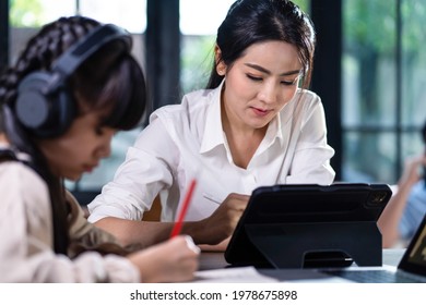 Homeschool Asian Little Young Girl Learning Online Class From School Teacher By Digital Remote Internet Meeting Due To Coronavirus Pandemic. Kid Looking Computer And Writing Note With Help From Mother