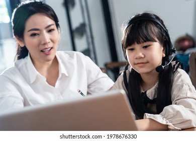 Homeschool Asian Little Young Girl Learning Online Class From School Teacher By Digital Remote Internet Meeting Due To Coronavirus Pandemic. Kid Looking Computer And Writing Note With Help From Mother