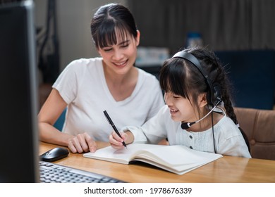 Homeschool Asian Little Young Girl Learning Online Class From School Teacher By Digital Remote Internet Meeting Due To Coronavirus Pandemic. Kid Looking Computer And Writing Note With Help From Mother