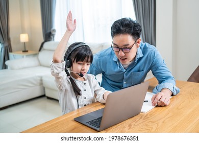 Homeschool Asian Little Young Girl Learning Online Class From School Teacher By Digital Remote Internet Meeting Due To Coronavirus Pandemic. Kid Looking Computer And Writing Note With Help From Father