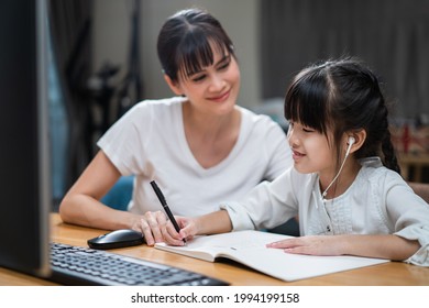 Homeschool Asian Little Young Girl Learning Online Class From School Teacher By Digital Remote Internet Meeting Due To Coronavirus Pandemic. Kid Looking Computer And Writing Note With Help From Mother