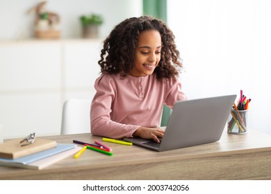 Homeschooling concept. Happy black girl studying at home with laptop, typing on keyboard, sitting at table in living room, having web lesson. Schoolgirl smiling and enjoying distance learning