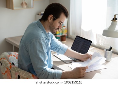 Homesourcing. Concentrated young man sit by laptop pc at home office doing paperwork reading document hardcopy. Remote worker employee analyzing statistic data correcting mistakes in printed report - Shutterstock ID 1902153118