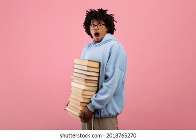 A Lot Of Homework. Terrified Black Teenage Student Standing With Huge Stack Of Books, Opening Mouth In Shock On Pink Background. Overwhelmed Teen Guy Preparing For College Entrance Or Graduation Exam