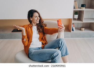 Homey caucasian girl has video phone call. Relaxed young woman is using airpods and cellphone at home. Wireless earphones using at online meeting. Technology using, leisure and communication concept.