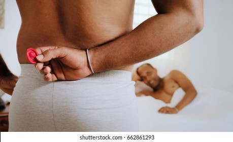 Homosexual Couple, Gay People, Same Sex Marriage Between Hispanic Men. Man In Bedroom Holding Condom For Safe Sex.