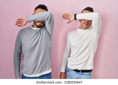 Homosexual Couple Standing Over Pink Background Covering Eyes With Arm, Looking Serious And Sad. Sightless, Hiding And Rejection Concept 