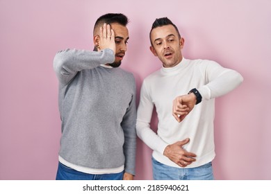 Homosexual Couple Standing Over Pink Background Looking At The Watch Time Worried, Afraid Of Getting Late 