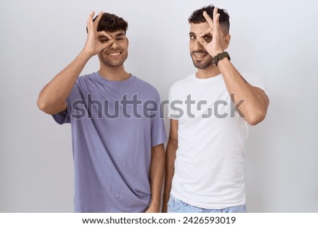 Homosexual gay couple standing over white background doing ok gesture with hand smiling, eye looking through fingers with happy face. 
