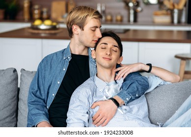 Homosexual guys. Loving gay couple, sitting at home, tenderly hugging and caressing each other, relaxing together on the sofa in the living room. Homosexual relationship concept