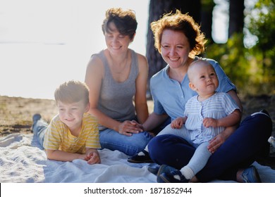 Homosexual Lesbian Family With Two Children, A Son And A Daughter. Two Moms And Kids At An Outdoor Picnic. Forest And Sea. Summer Day At Sunset.