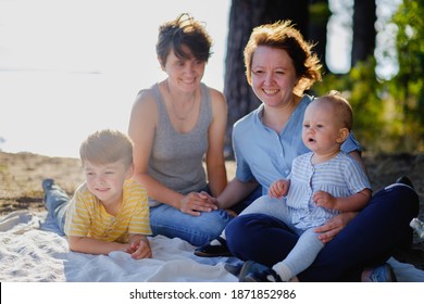 Homosexual Lesbian Family With Two Children, A Son And A Daughter. Two Moms And Kids At An Outdoor Picnic. Forest And Sea. Summer Day At Sunset.