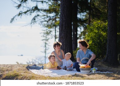 Homosexual Lesbian Family With Two Children, A Son And A Daughter. Two Moms And Kids At An Outdoor Picnic. Forest And Sea. Summer Day At Sunset.