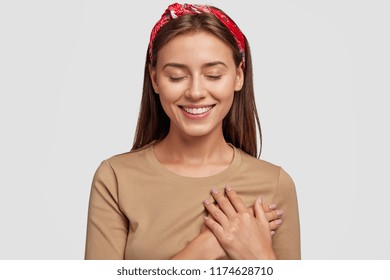 Honest kind hearted woman keeps both palms on chest, expresses kindness, keeps eyes shut, dressed in casual outfit, isolated over white background. People, feeling and body language concept.