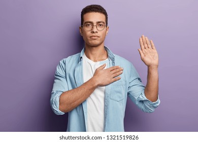 Honest serious hipster guy swears to do something, keeps hand on chest, gestures with palm, promises something, wears spectacles and denim shirt, swears indoor, isolated over purple background