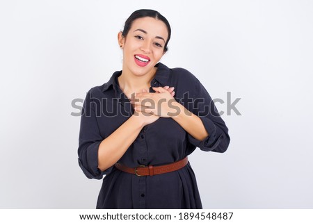 Honest young beautiful Arab woman wearing gray dress against white studio background keeps hands on chest, touched by compliment or makes promise, looks at camera with great pleasure.