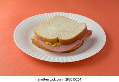 A Honey Ham Sandwich With Mustard On White Bread Upon A Paper Plate And Orange Table Top.