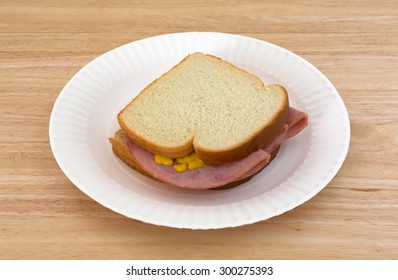 A Honey Ham Sandwich With Mustard On White Bread Upon A Paper Plate And Wood Table Top.