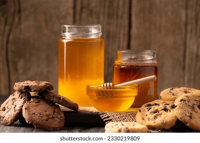 Honey in open jars and in a bowl, dipper and homemade cookies with chocolate chips on a wooden background. Homemade cakes and honey for breakfast. Composition of craft chocolate cookies and honey. - Powered by Shutterstock