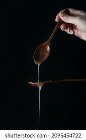 Honey Is Poured Onto A Wooden Spoon. Black Background. Transparent Honey Drips From A Spoon. The Hand Holds A Wooden Spoon Dripping With Honey