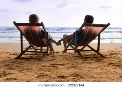 Honeymoon at sunset tropical beach in Thailand. Romantic couple are sitting in deck chairs and holding their hands.