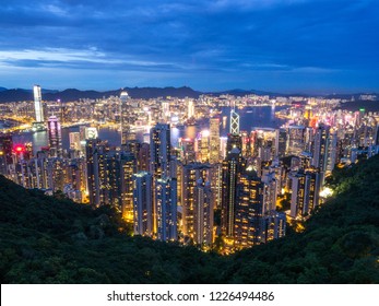 Hong Kong Skyline At Night From The Famous Victoria Point At Daytime. Green Forest And River Surrounded By Towering Sky Scrapers Which Is Densly Populated. Deep Blue Sky And Illuminated Tall Buildings