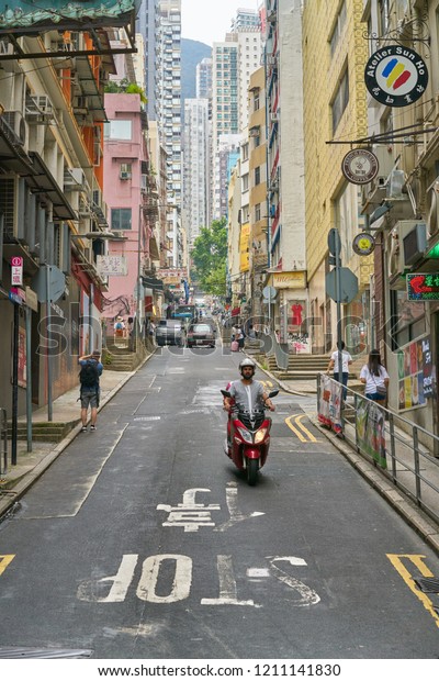 HONGKONG - OCTOBER 1, 2018: In a morning street near\
Sheung-wan, a motorcycle rider is driving by. This is a typical HK\
street, narrow road surrounded by buildings of various stores and\
restaurants. 