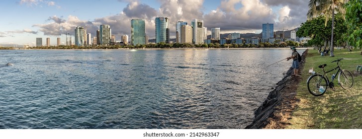 Honolulu, Hawaii - circa February 2022: A person fishes at Ala Moana harbor. Panorama of the city skyline beyond.