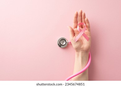 Honor Breast Cancer Awareness Month with this top view image of female hand holding pink ribbon and stethoscope on pastel pink isolated background with copyspace available for text or ads