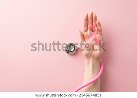 Honor Breast Cancer Awareness Month with this top view image of female hand holding pink ribbon and stethoscope on pastel pink isolated background with copyspace available for text or ads