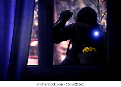 Hooded Burglar Forcing The Window Frame - Silhouette Of Thief With Screwdriver And Flashlight Is Breaking Into The Apartment - Concept Of Intrusion And Danger View From Indoor Dark Shaded Image