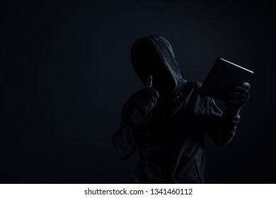 Hooded computer hacker with obscured face using digital tablet in cybercrime and cybersecurity concept, low key with selective focus - Shutterstock ID 1341460112