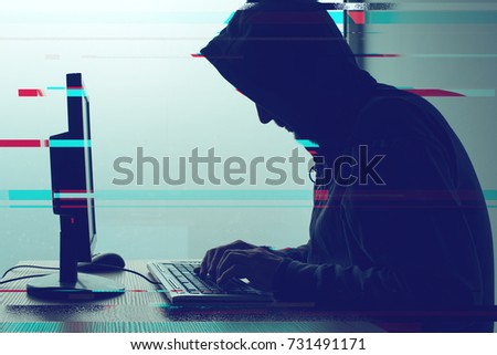 Hooded computer hacker working on desktop PC computer. P2P and piracy, computer virus and trojans, phishing, malware and ransomware concept with glitch effect.