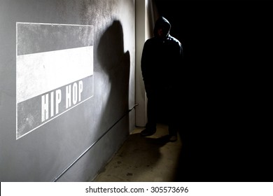 Hooded Hip Hop Rapper Next To Wall Graffiti With Copy Space.  The Man Is Standing In A Dark Alley And A Spray Painted Concrete Wall.