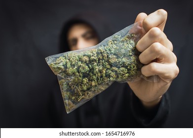 Hooded Man Holding A Big Bag Of Weed Front His Head On Black Background. Drug Trafficking Concept, Buy Or Sell Marijuana.