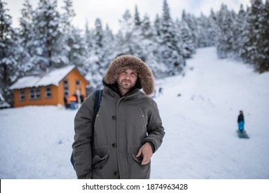 Hooded Man Standing With A Beautiful Snowy Forest And Small Cabin As Background. Portrait Of Happy Man Surrounded By Snowy Pine Trees. Adventurous Winter Holiday