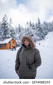 Hooded Man Standing With A Beautiful Snowy Forest And Small Cabin As Background. Portrait Of Happy Man Surrounded By Snowy Pine Trees. Adventurous Winter Holiday