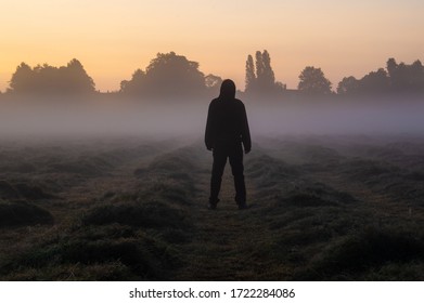 A hooded man standing in a misty field back to camera looking into the sky, Just before sunrise. - Powered by Shutterstock