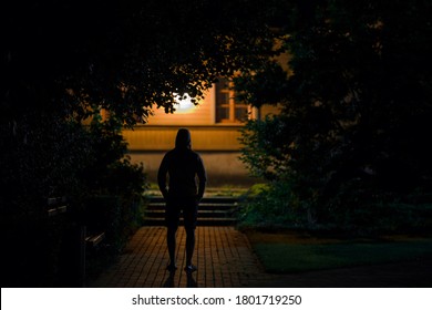 Hooded Man Standing On Sidewalk At Dark Park In Summer Black Night. Scary Moment And Gloomy Atmosphere. Thief Watching House. Back View.