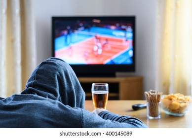 Hooded man watching TV (volleyball match) in living room with alcohol and snacks - stock photo
